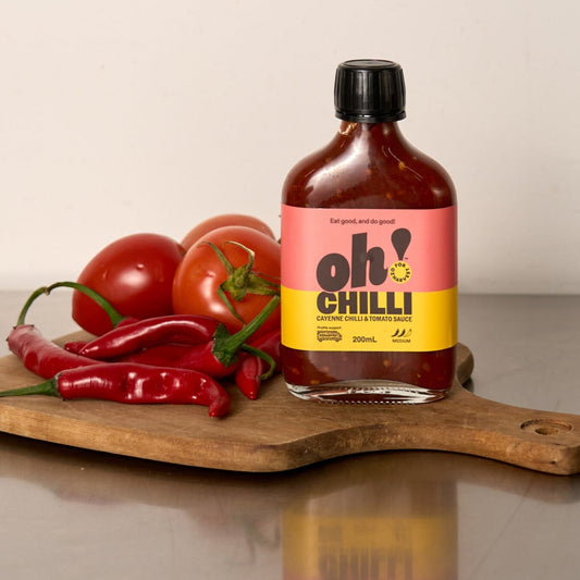 Bottle of Oh! Chilli sauce on a wooden cutting board with tomatoes and red peppers.