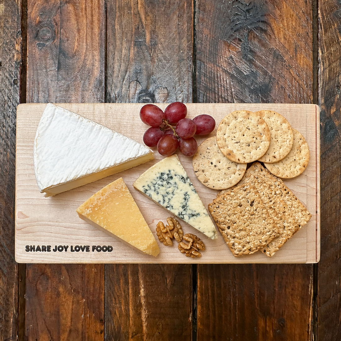 A wooden charcuterie board with cheese, crackers, grapes, and walnuts, photographed from above on a dark rustic wooden table. The board has the text 'SHARE JOY LOVE FOOD' printed on the bottom left.