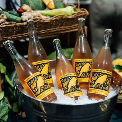 Bottles of a beverage in a metal bucket with ice, surrounded by vegetables.