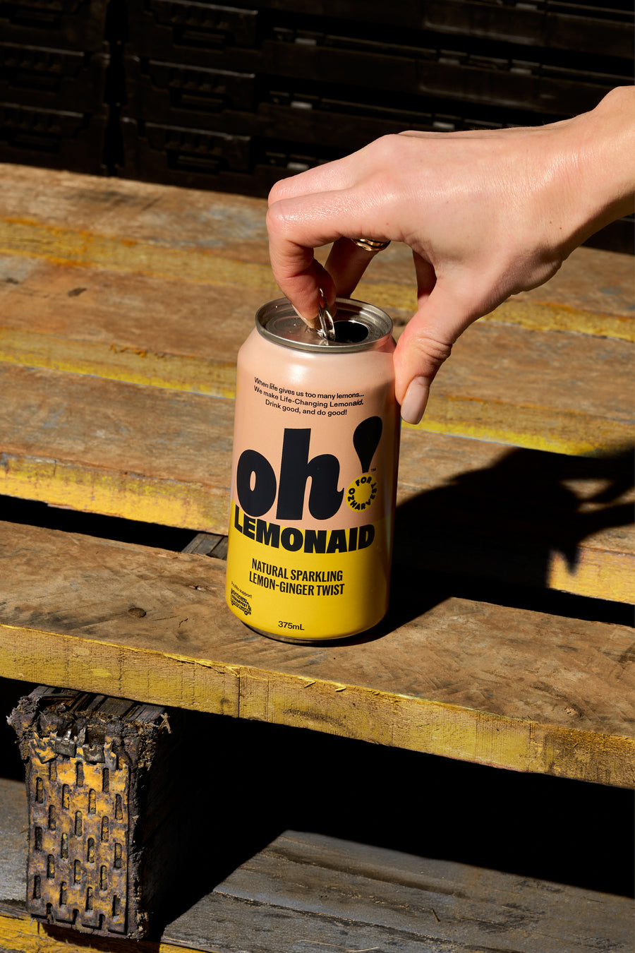 A hand cracking open a can of Oh! Lemonaid on a timber pallet