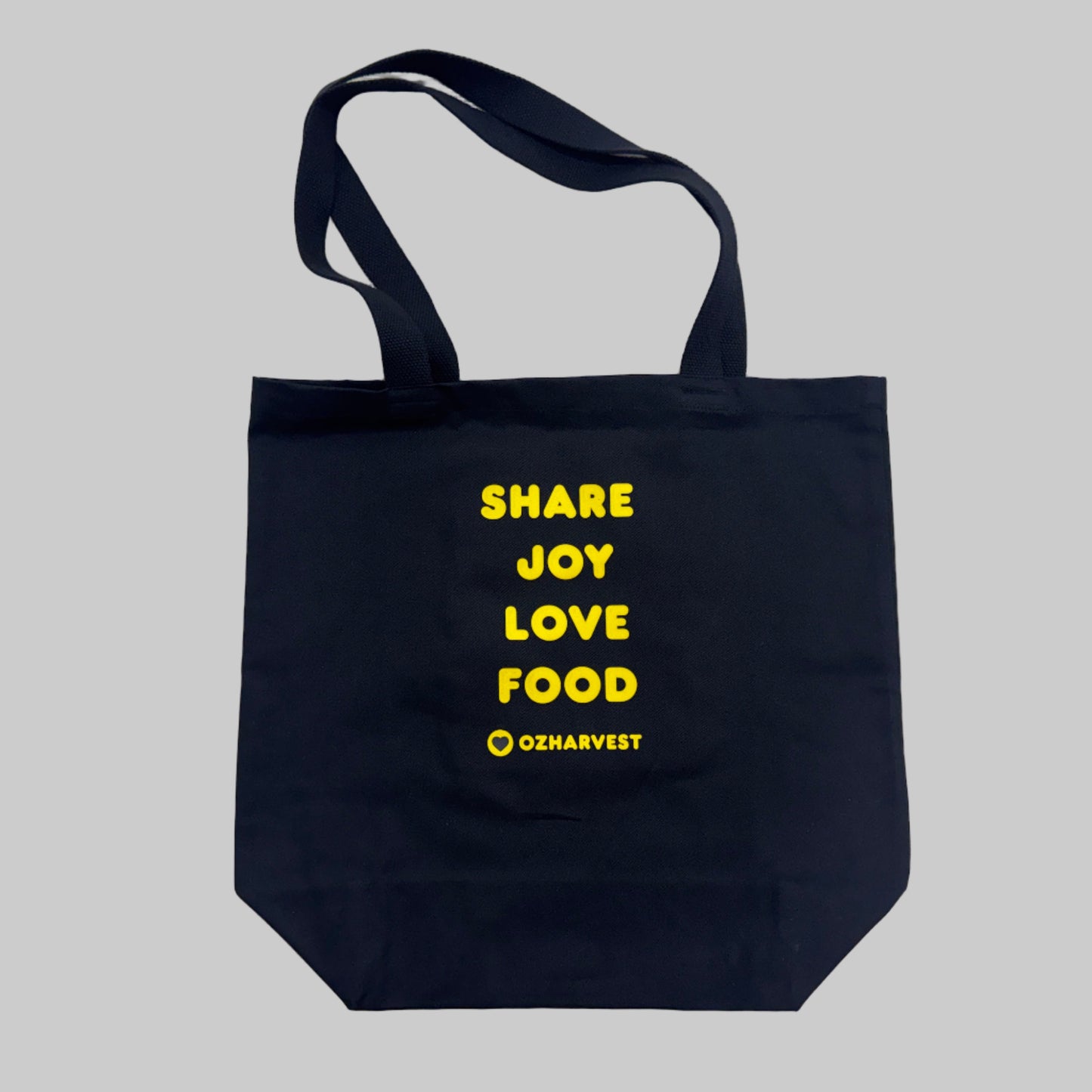 Black tote bag with yellow text 'SHARE JOY LOVE FOOD' and OZharvest logo on a light gray background