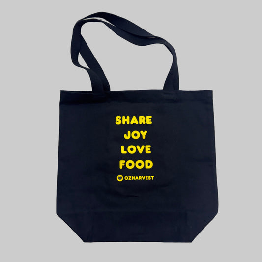 Black tote bag with yellow text 'SHARE JOY LOVE FOOD' and OZharvest logo on a light gray background